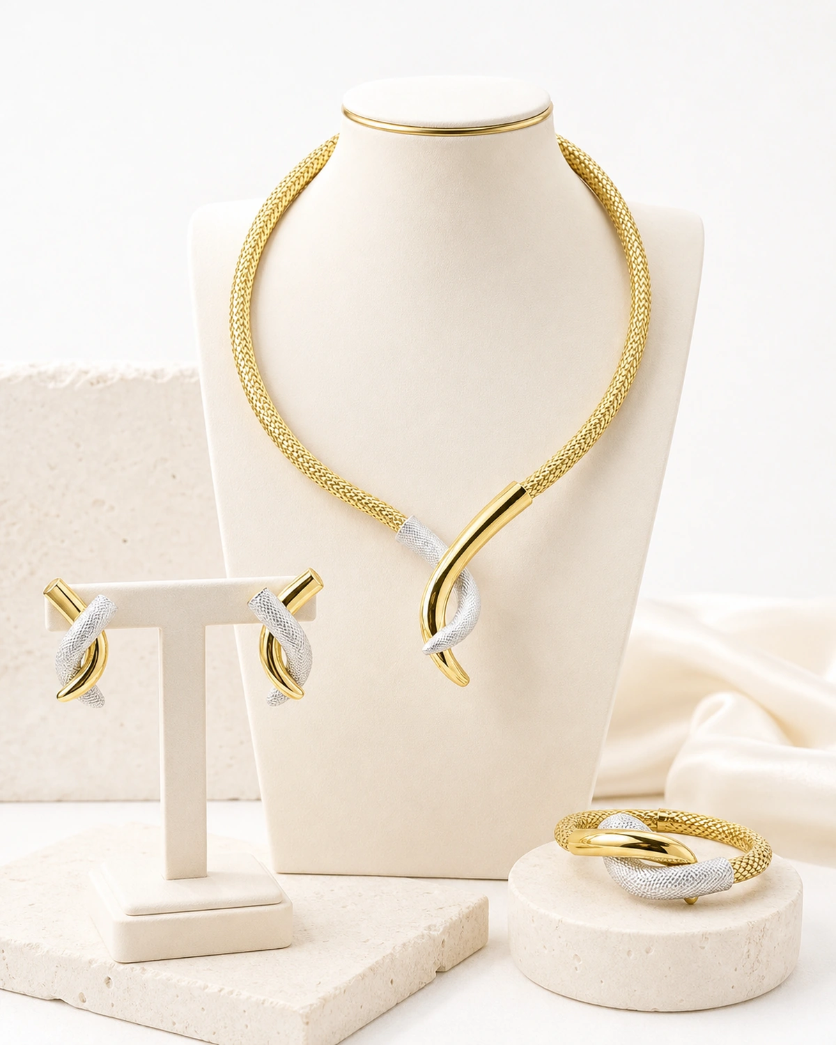 Catalogue hero of the complete Aurelia set with the rounded woven gold necklace, asymmetric gold and textured white-silver centerpiece, two distinct earrings and matching bracelet on a warm ivory display.