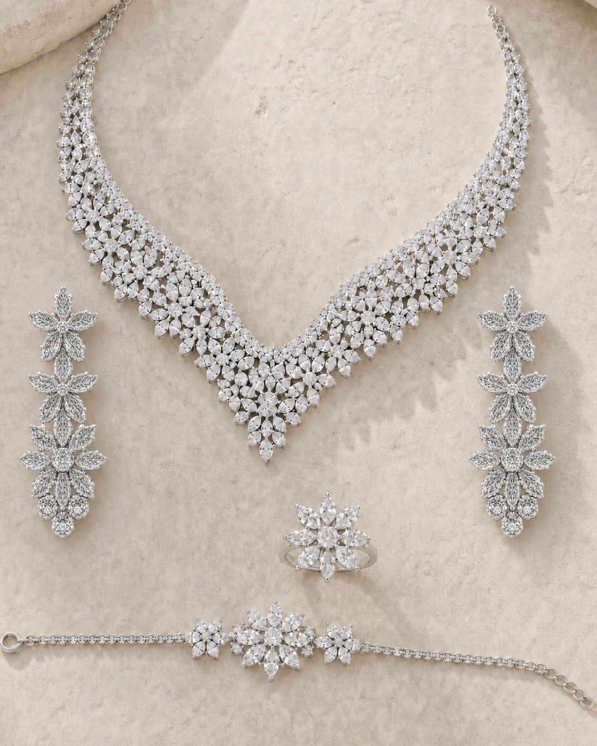 Regina catalogue hero showing the complete silver-tone floral V-bib necklace, matching long floral-drop earrings, compact starburst ring and fine three-flower-centre bracelet on Pale Stone.