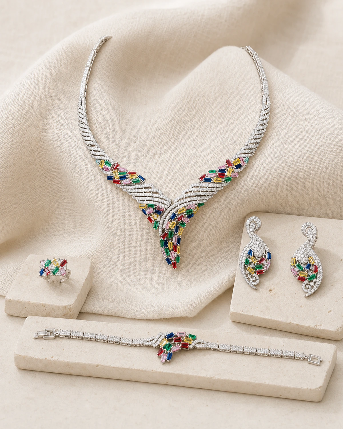 Catalogue hero of the complete five-piece Splendor set with one braided multi-row silver-tone V necklace, two curled drop earrings, one clustered ring and one central-cluster bracelet on warm-ivory linen and pale stone.