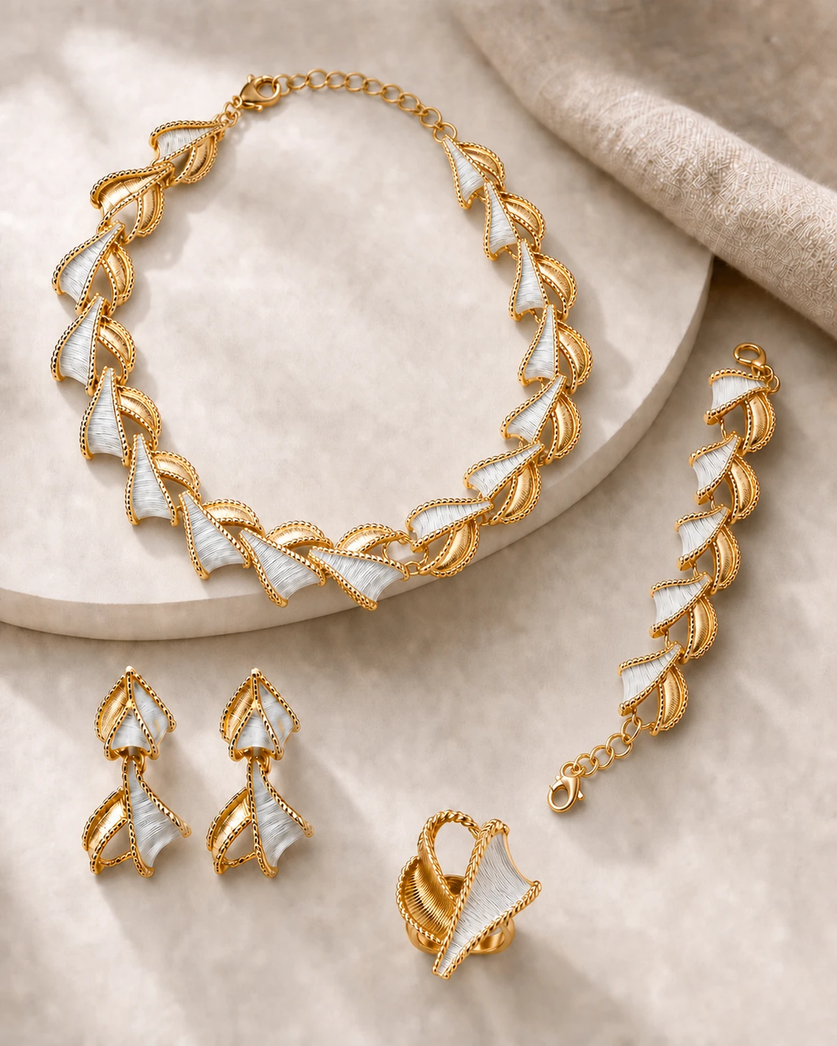 Catalogue hero of the complete Vesper set with one sculptural necklace, two matching two-tier earrings, one crossed fan-and-leaf ring and one repeating bracelet on pale stone and warm-taupe linen.
